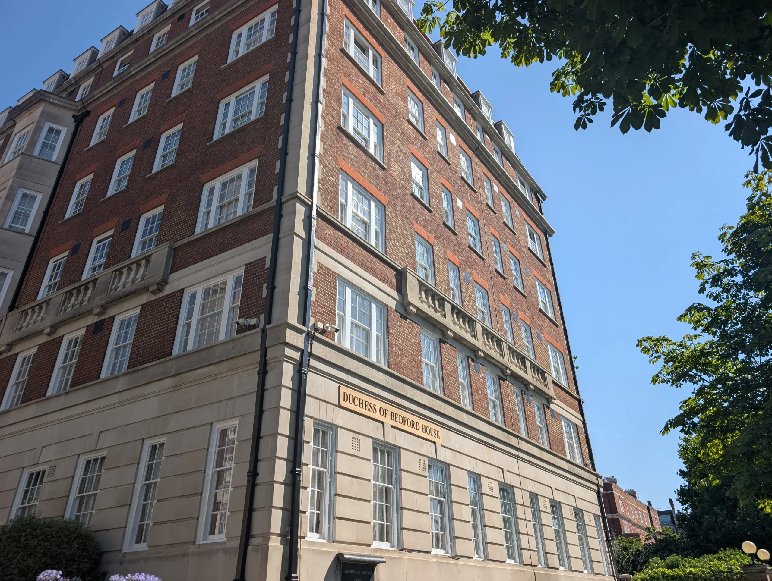 Alt text option Exterior view of a multi storey brick and stone building with windows, and a sign showing the name Duchess of Bedford House on the facade under a clear blue sky.