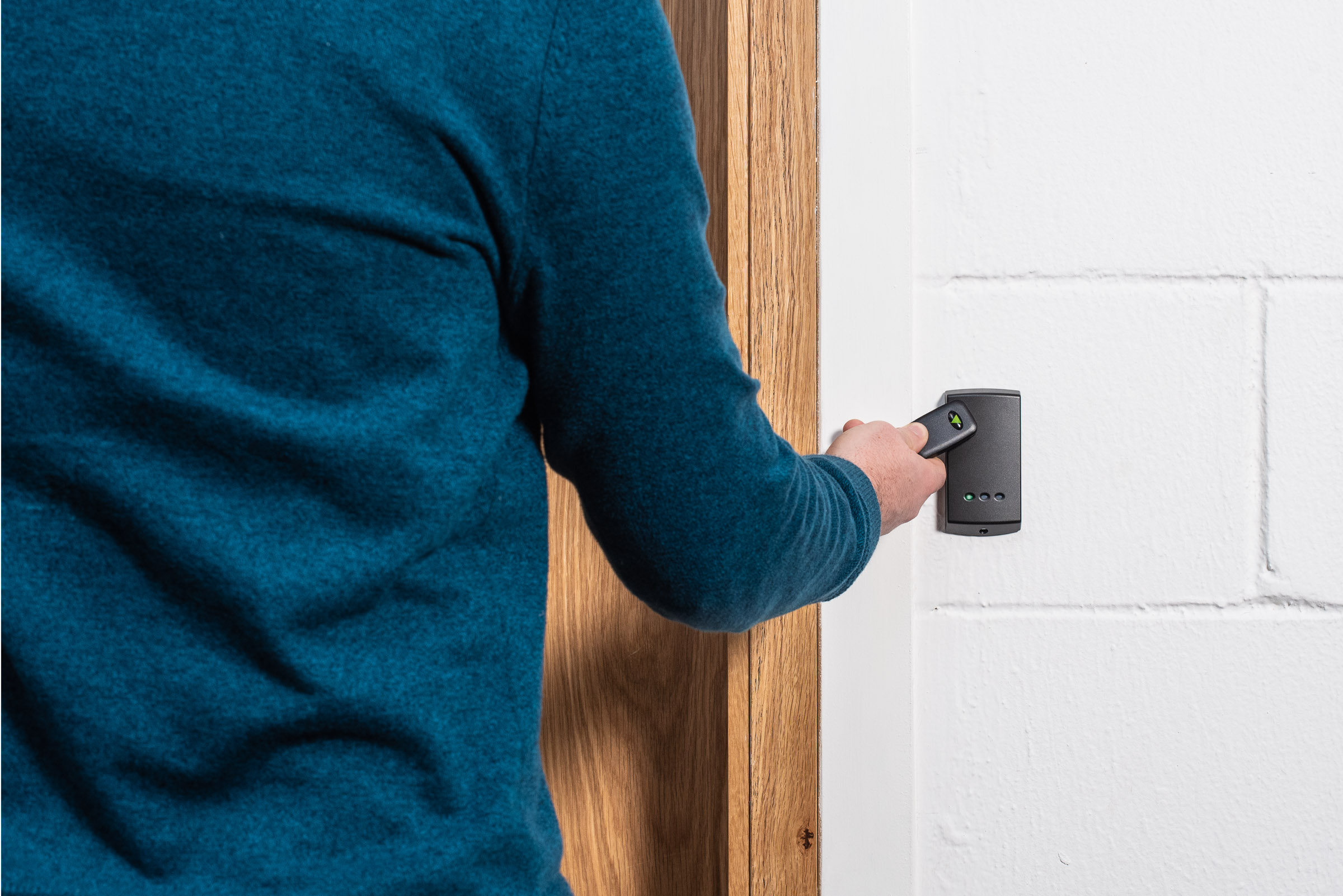 Person presenting a contactless key fob to a wall mounted access control reader next to a wooden door, with indicator lights visible on the reader.