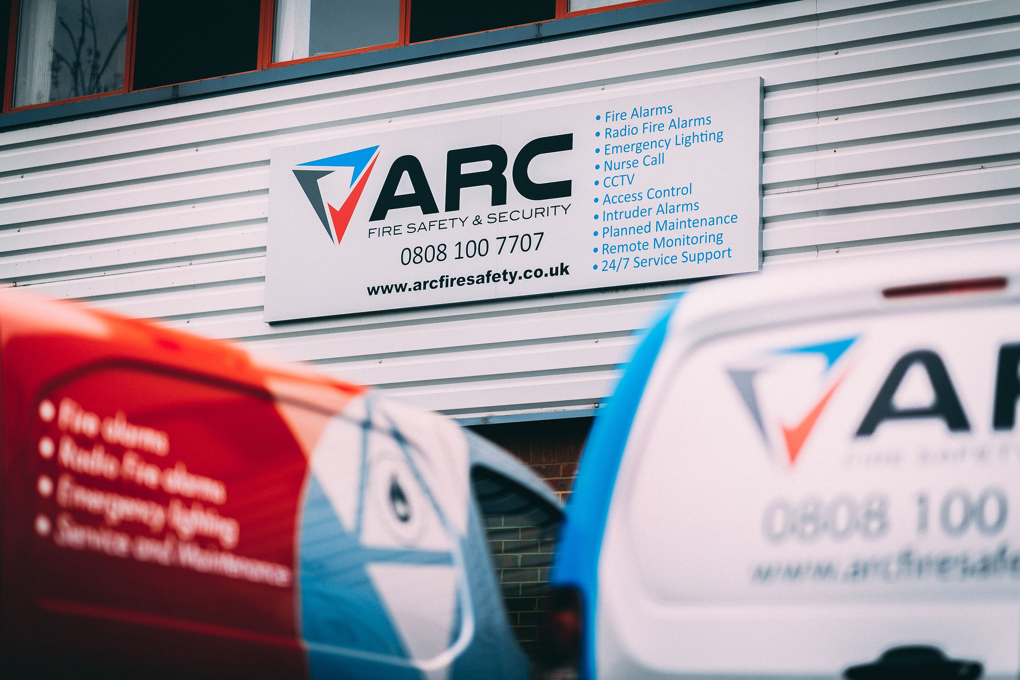 Exterior view of an ARC Fire Safety and Security building sign mounted on metal cladding, listing services such as fire alarms, CCTV, access control, and 24 7 support, with branded service vans partially visible in the foreground.