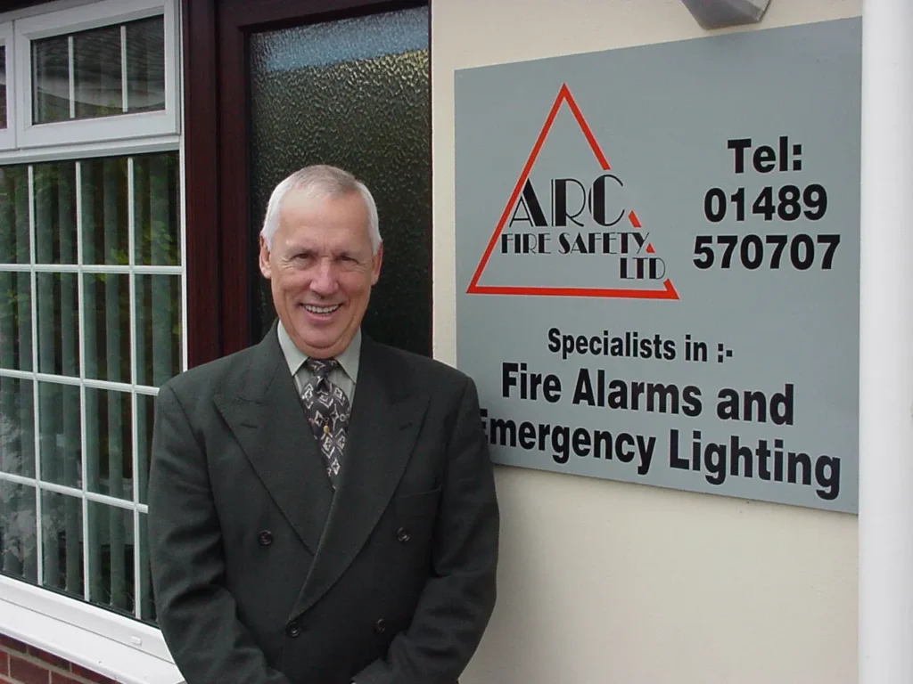 Person standing outside a building beside a wall mounted sign reading ARC Fire Safety Ltd, Specialists in Fire Alarms and Emergency Lighting, with a telephone number displayed on the sign.