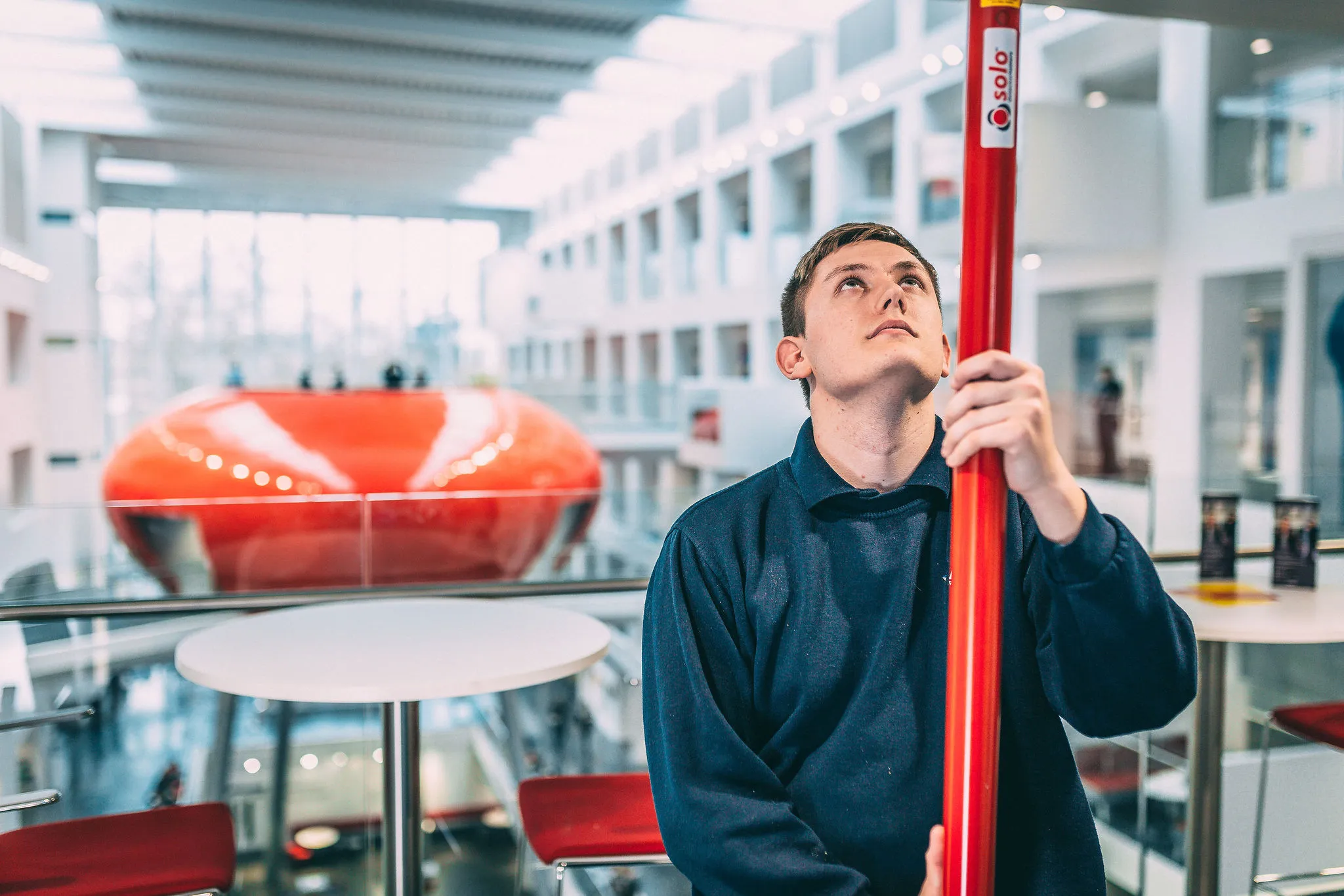 Person standing indoors holding a red safety pole, positioned beside a glass railing in a large modern atrium with multiple floors, tables, and seating visible in the background.