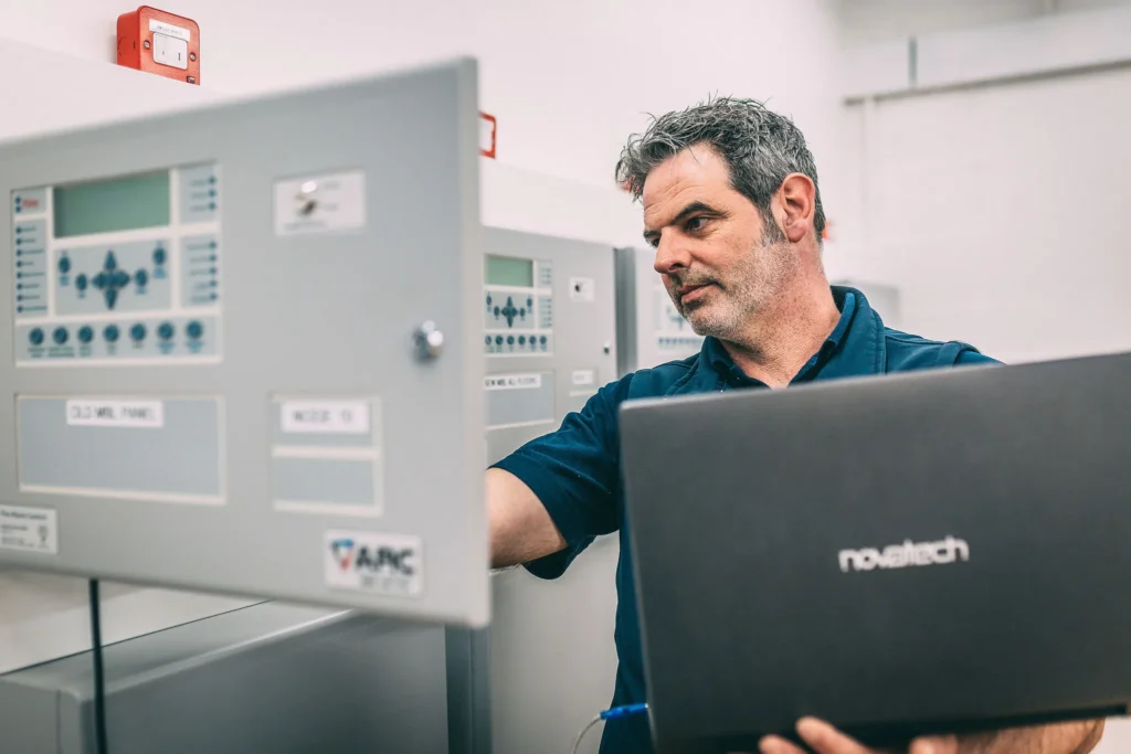 Person working in a plant room using a laptop beside wall mounted fire alarm control panels, with multiple control units and red alarm devices visible in the background.