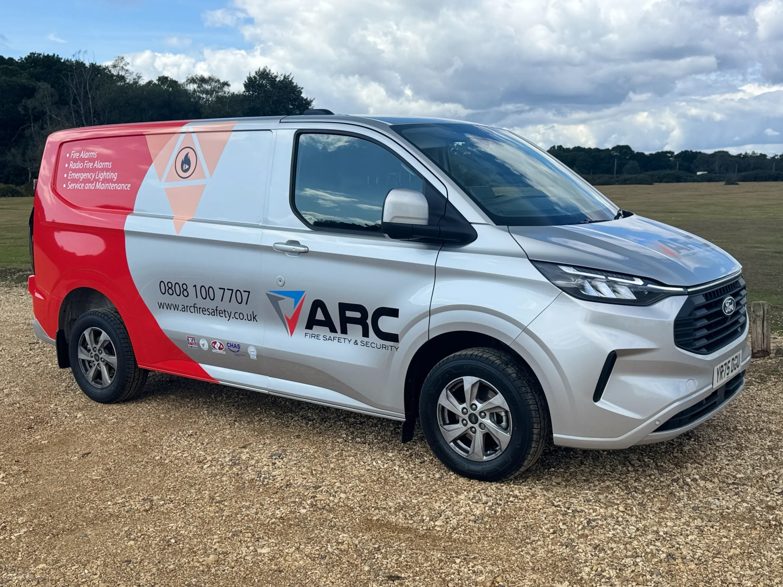 ARC Fire Safety and Security branded service van parked on a gravel surface in an open rural area, shown from the side with red and grey graphics and contact details visible.