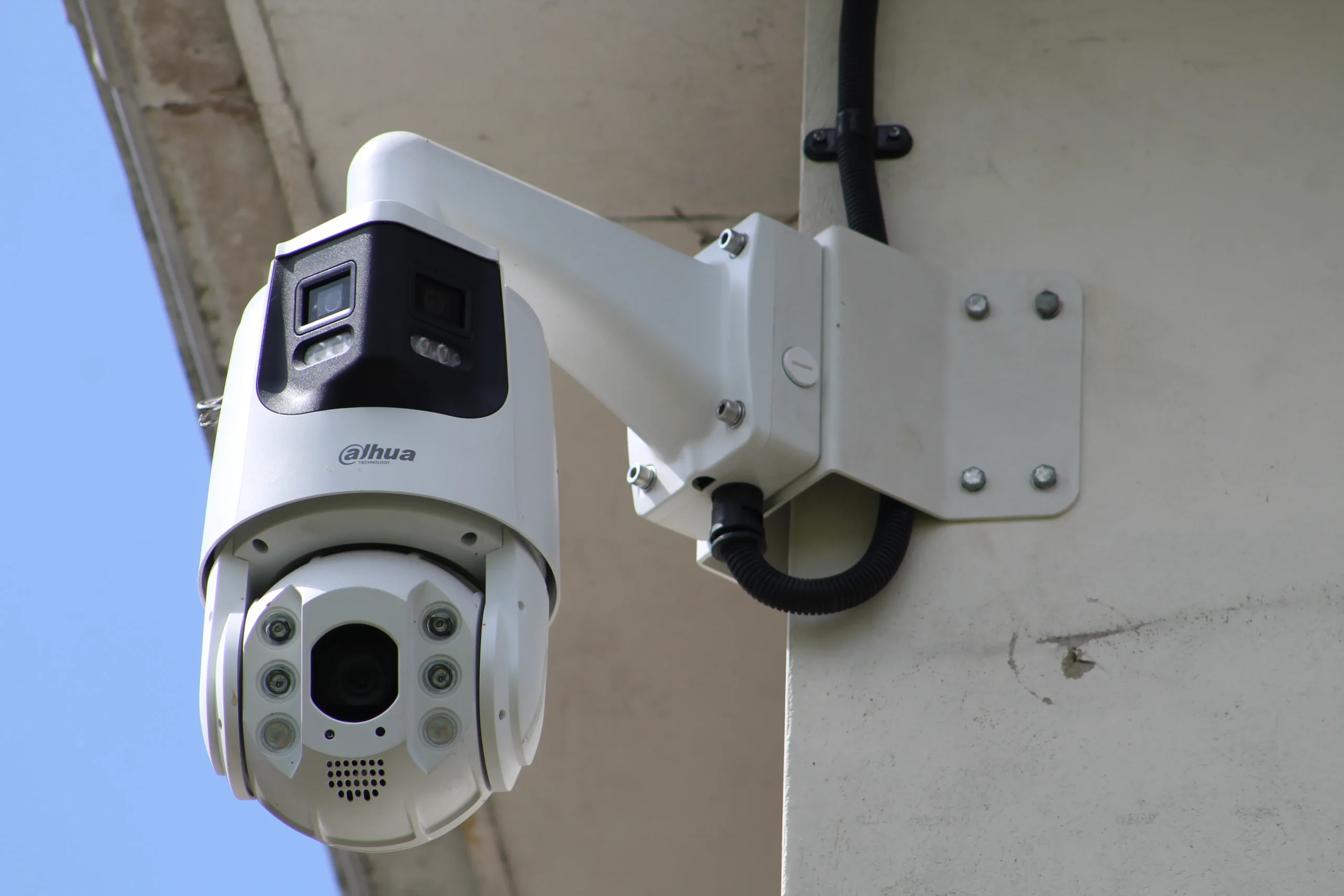 External PTZ security camera attached to a building corner using a white metal bracket, with visible cabling and a multi lens camera head positioned beneath the housing.