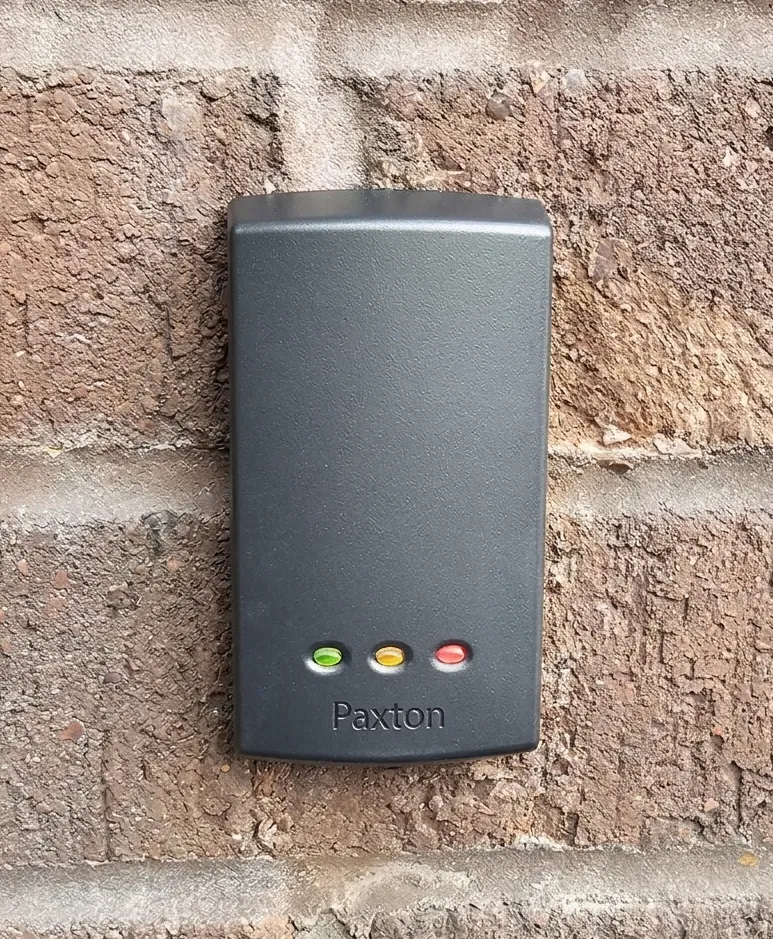 Paxton access control card reader mounted on a brick wall, featuring three status indicator lights in green, amber, and red.