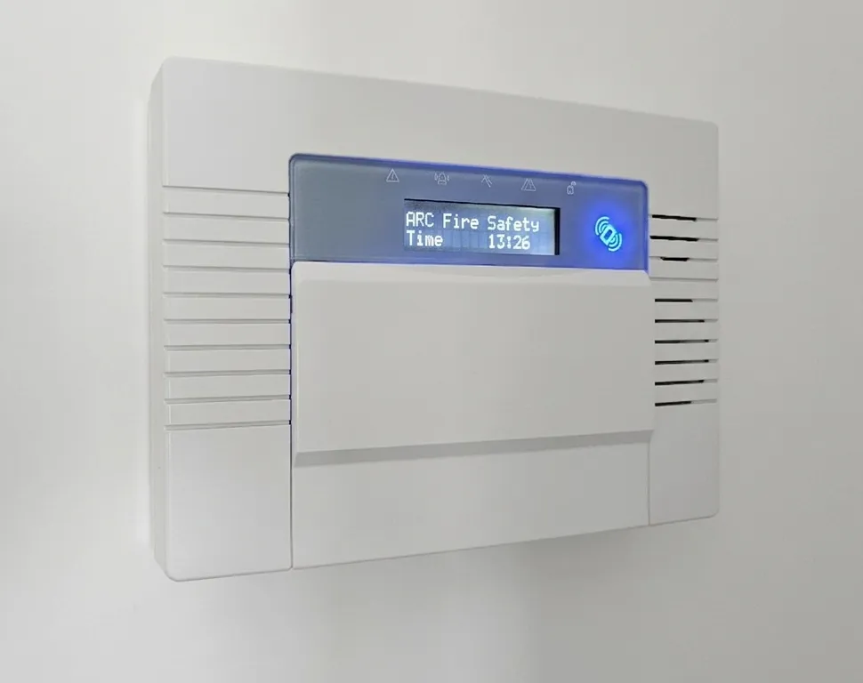 Wall mounted intruder alarm control panel with a digital display reading ARC Fire Safety and the time, featuring a blue illuminated indicator and a white casing.