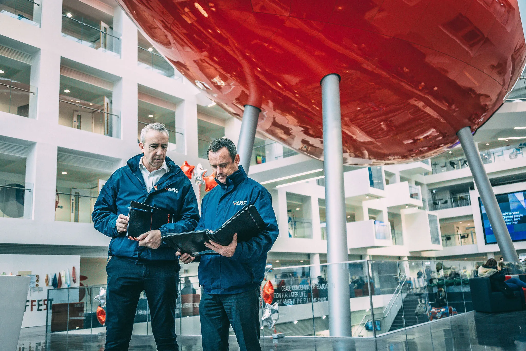 Two people wearing ARC branded jackets standing inside a large modern atrium, reviewing documents together beneath a suspended red sculptural structure, with multiple floors and glass walkways visible around them.