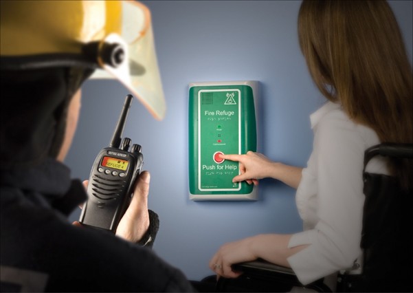 Close up of a fire refuge emergency call point mounted on a wall, showing a hand pressing the Push for Help button, with a handheld radio and protective helmet visible nearby.