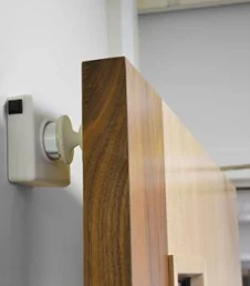 Close up of a door fitted with a wall mounted access control component, showing a wooden door edge positioned next to a small rectangular reader or sensor unit.
