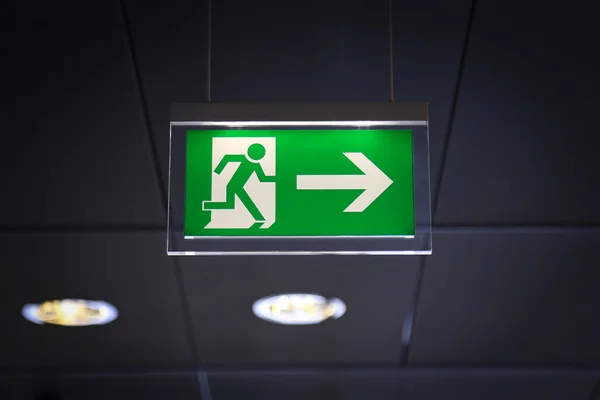 Illuminated green emergency exit sign suspended from a ceiling, displaying a running figure pictogram and a right pointing arrow indicating the direction of exit.