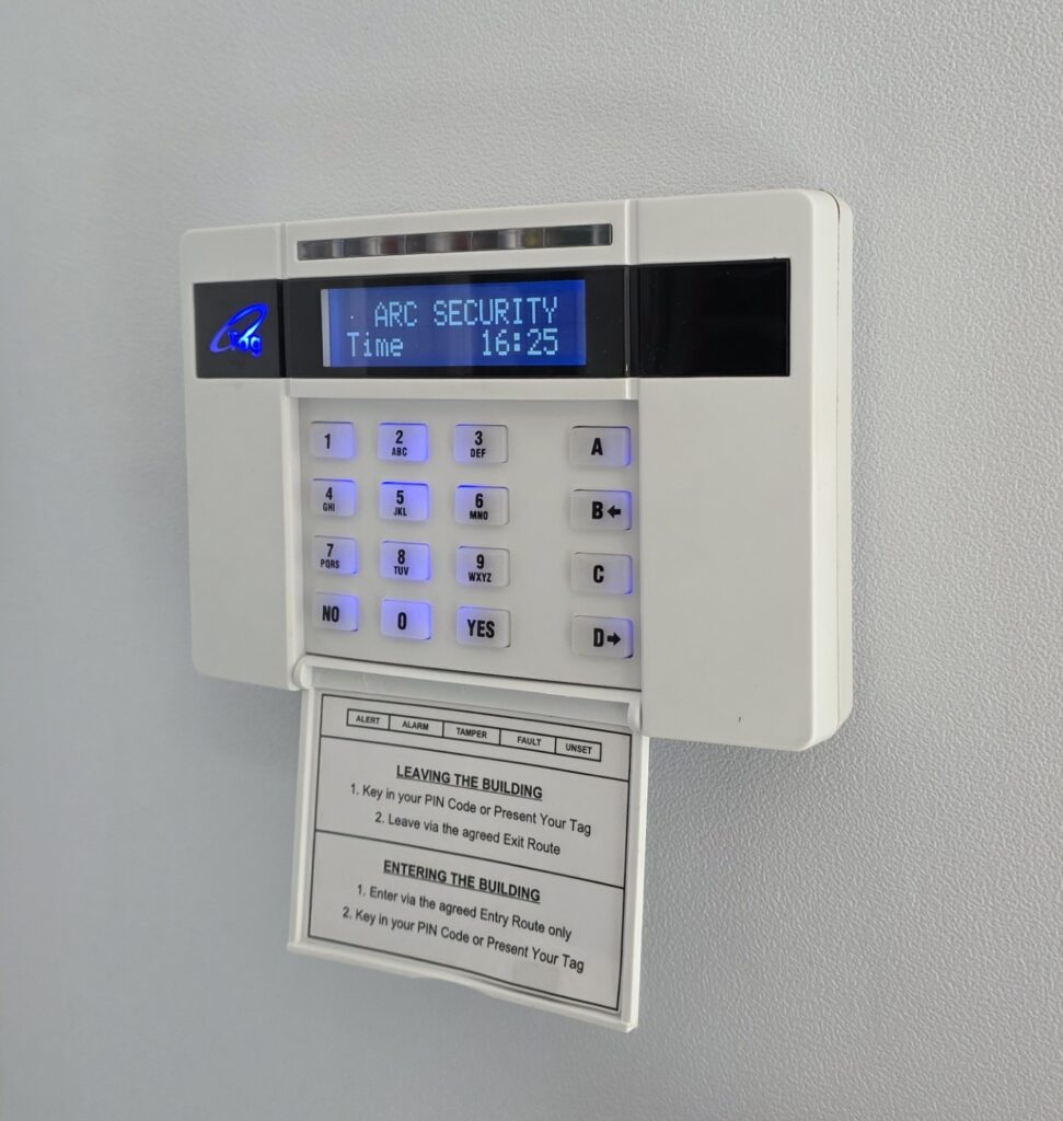 Wall mounted security alarm keypad with illuminated numeric buttons, digital display showing system status and time, and an instruction card positioned below.