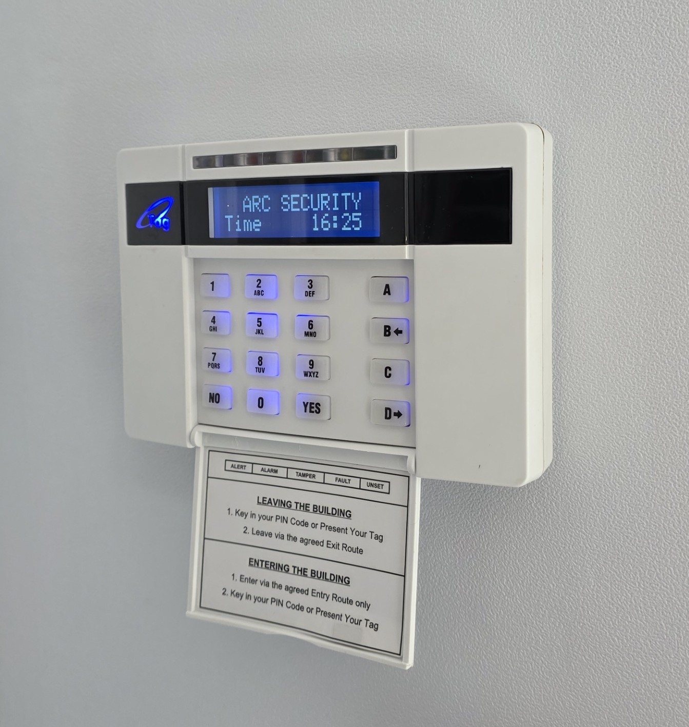 Wall mounted security alarm keypad with illuminated numeric buttons, digital display showing system status and time, and an instruction card positioned below.
