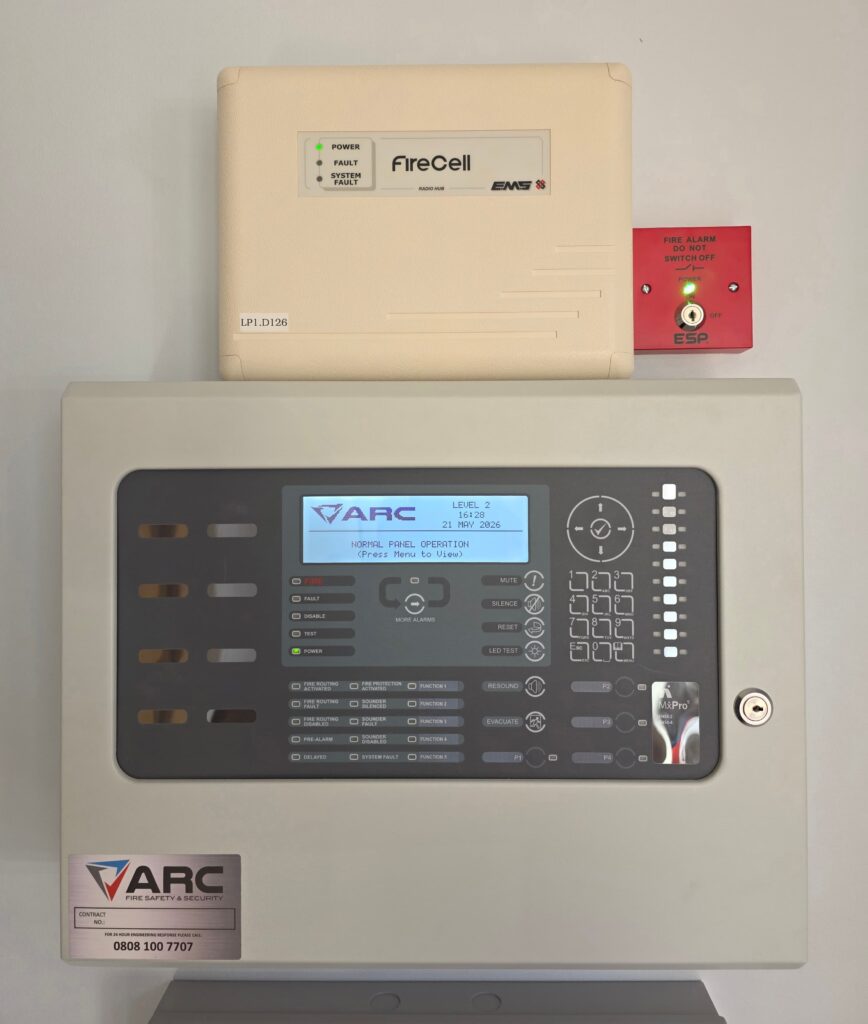 Wall mounted fire alarm control panel with digital display, keypad, and status indicators, with a FireCell wireless unit and a red fire alarm isolation switch mounted above.